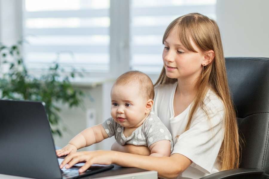 A young girl sitting at a desk with a baby on her lap, both looking at a laptop screen, illustrating early exposure to screen time.