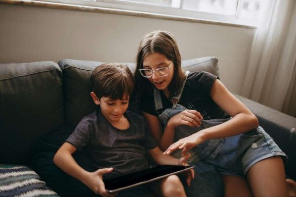 A boy and a girl sitting together on a gray sofa sharing a tablet, engaged in screen time during a relaxed afternoon at home.