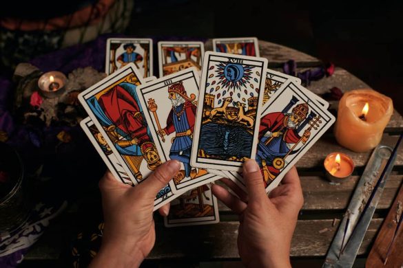Two hands holding a fan of traditional tarot cards over a wooden table decorated with lit candles