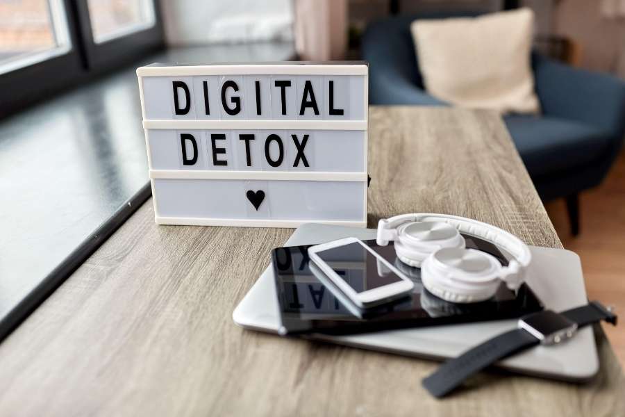 A lightbox sign displaying "DIGITAL DETOX" with a heart symbol sits on a wooden desk, surrounded by a stacked smartphone, tablet, smartwatch, and headphones that have been put away.
